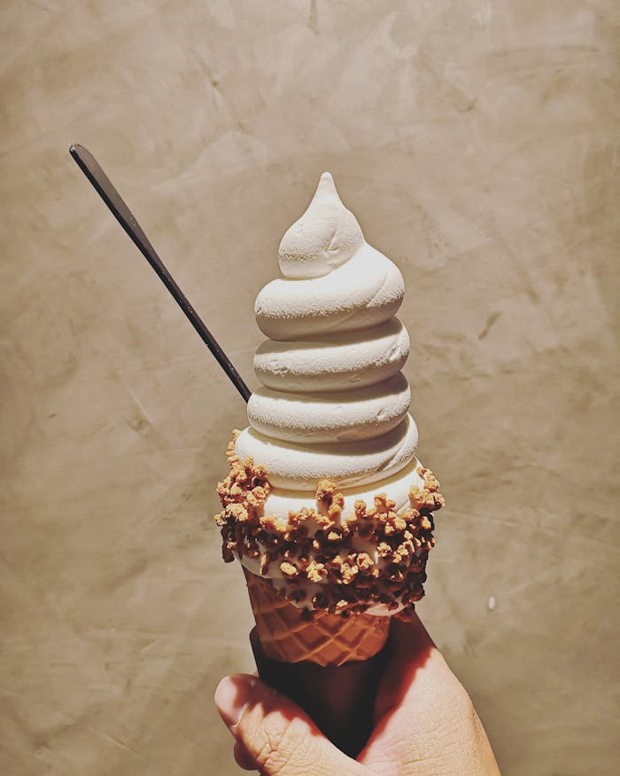 Close-up of a vanilla ice cream cone with textured waffle cone and nut toppings held by a hand.